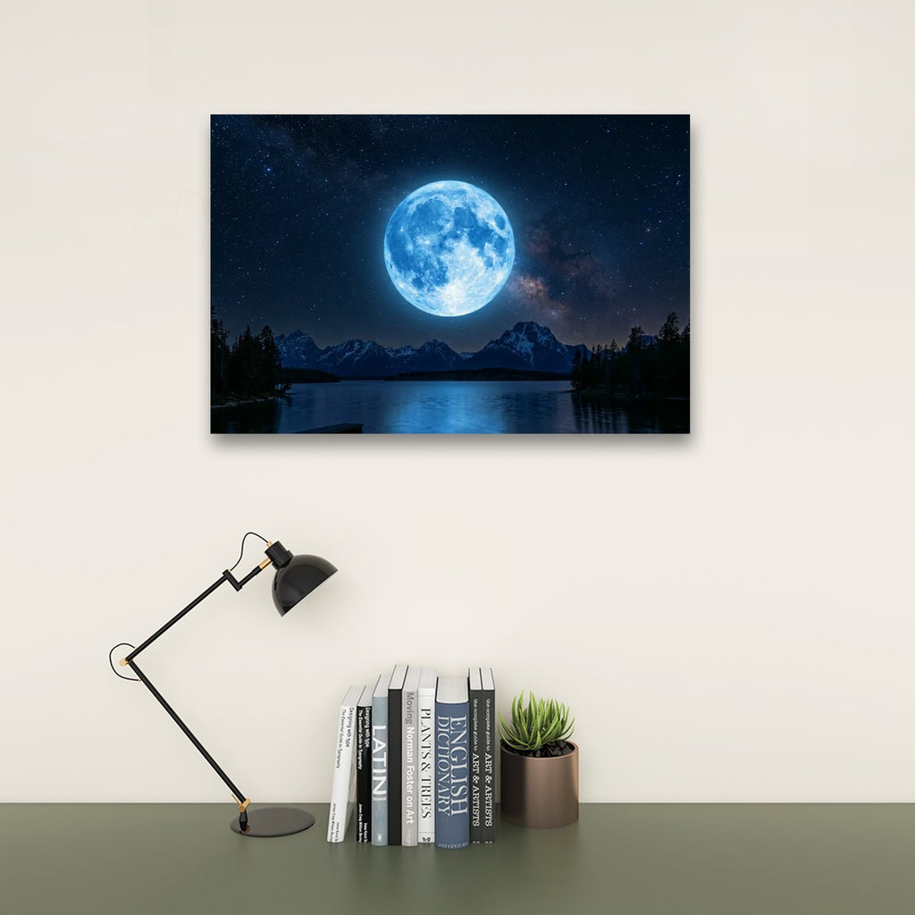 A rectangular wall art print showing a glowing blue full moon over a dark mountain lake at night, displayed above a desk with a black lamp, books, and a small succulent on a light wall.