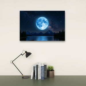 A rectangular wall art print showing a glowing blue full moon over a dark mountain lake at night, displayed above a desk with a black lamp, books, and a small succulent on a light wall.