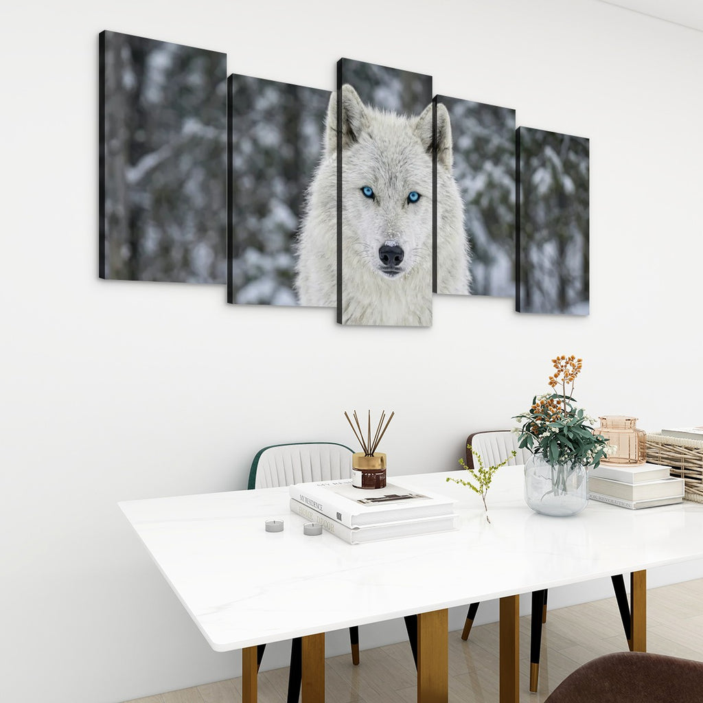 A five-panel canvas wall art set depicting a close-up of a white wolf with bright blue eyes in a snowy forest, displayed on a white wall above a modern dining table with books and a vase.
