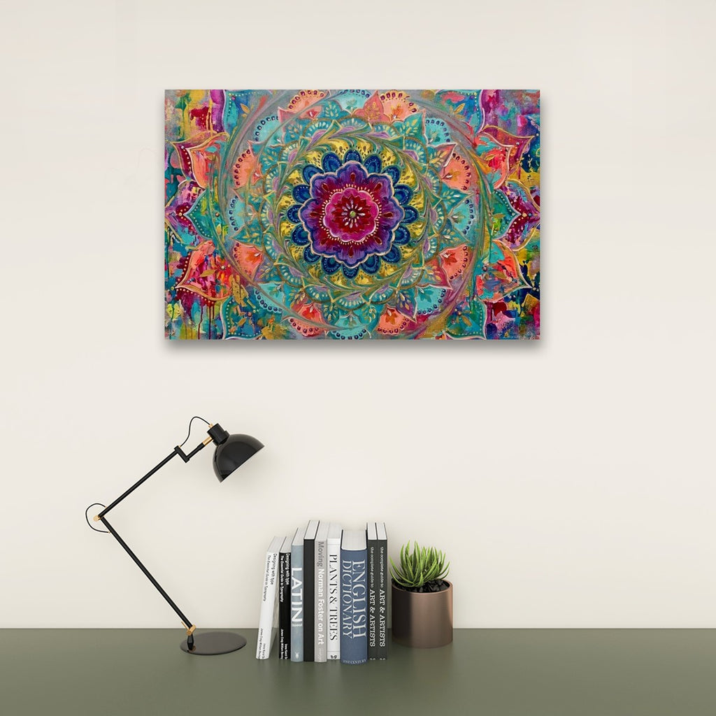 A colorful square wall art print featuring a vibrant mandala design, displayed on a light beige wall above a dark green desk with a black desk lamp, books, and a small potted succulent.