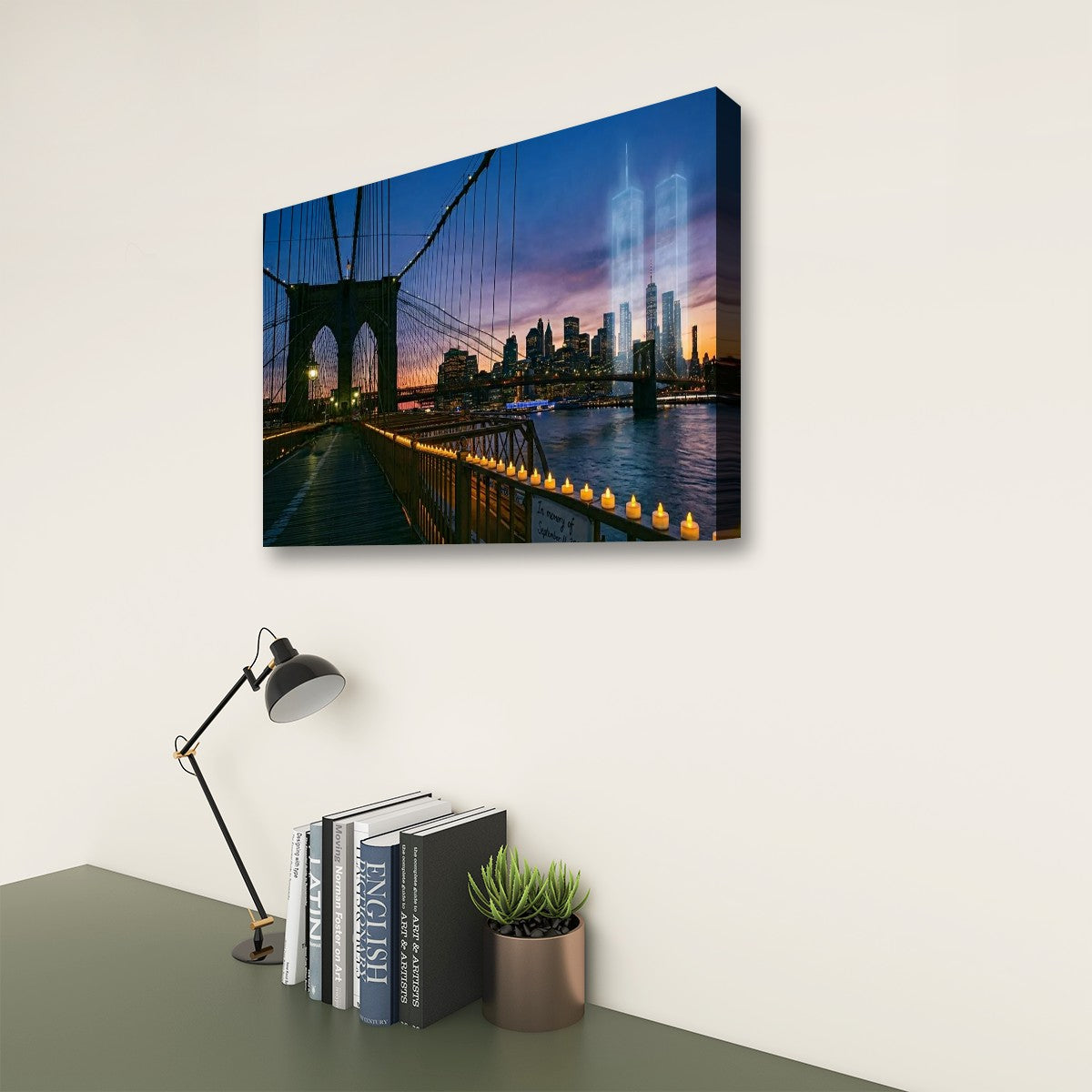 A rectangular wall art print showing a city skyline at dusk with a suspension bridge and illuminated buildings, displayed on a light wall above a desk with a lamp, books, and a small plant.