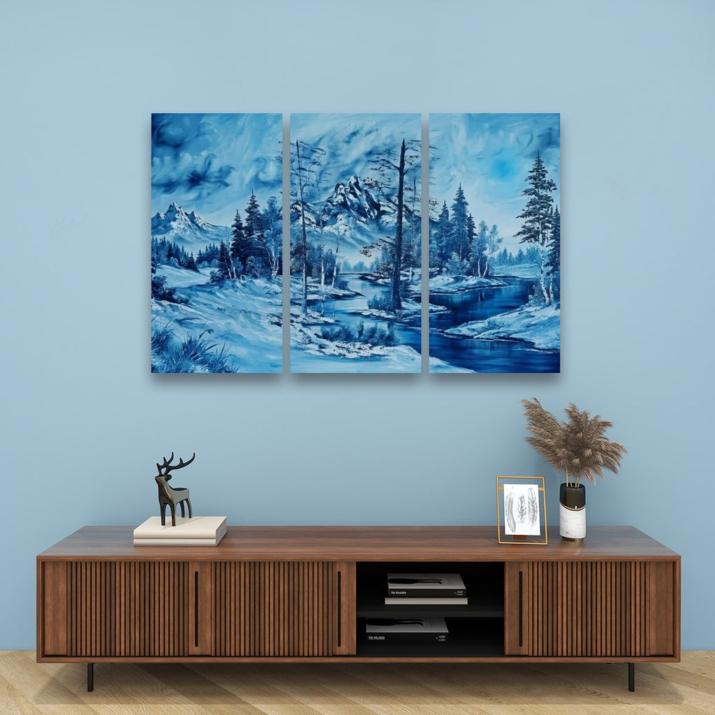 A three-panel canvas wall art set depicting a blue-toned winter landscape with snowy mountains, evergreen trees, and a river, displayed above a wooden media console against a light blue wall.