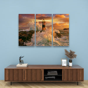 A three-panel canvas wall art set depicting a dramatic lighthouse scene with crashing waves and a golden sunset, displayed on a light blue wall above a wooden console table with decorative items.