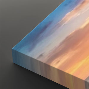 A close-up of a rectangular canvas or print showing an abstract landscape-like scene with blue sky, soft white clouds, and warm peach-orange sunlight, photographed against a dark gray background.