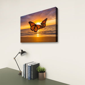 A rectangular wall art print or canvas showing two orange butterflies over a beach sunset, displayed on a light wall above a desk with books, a lamp, and a small plant.