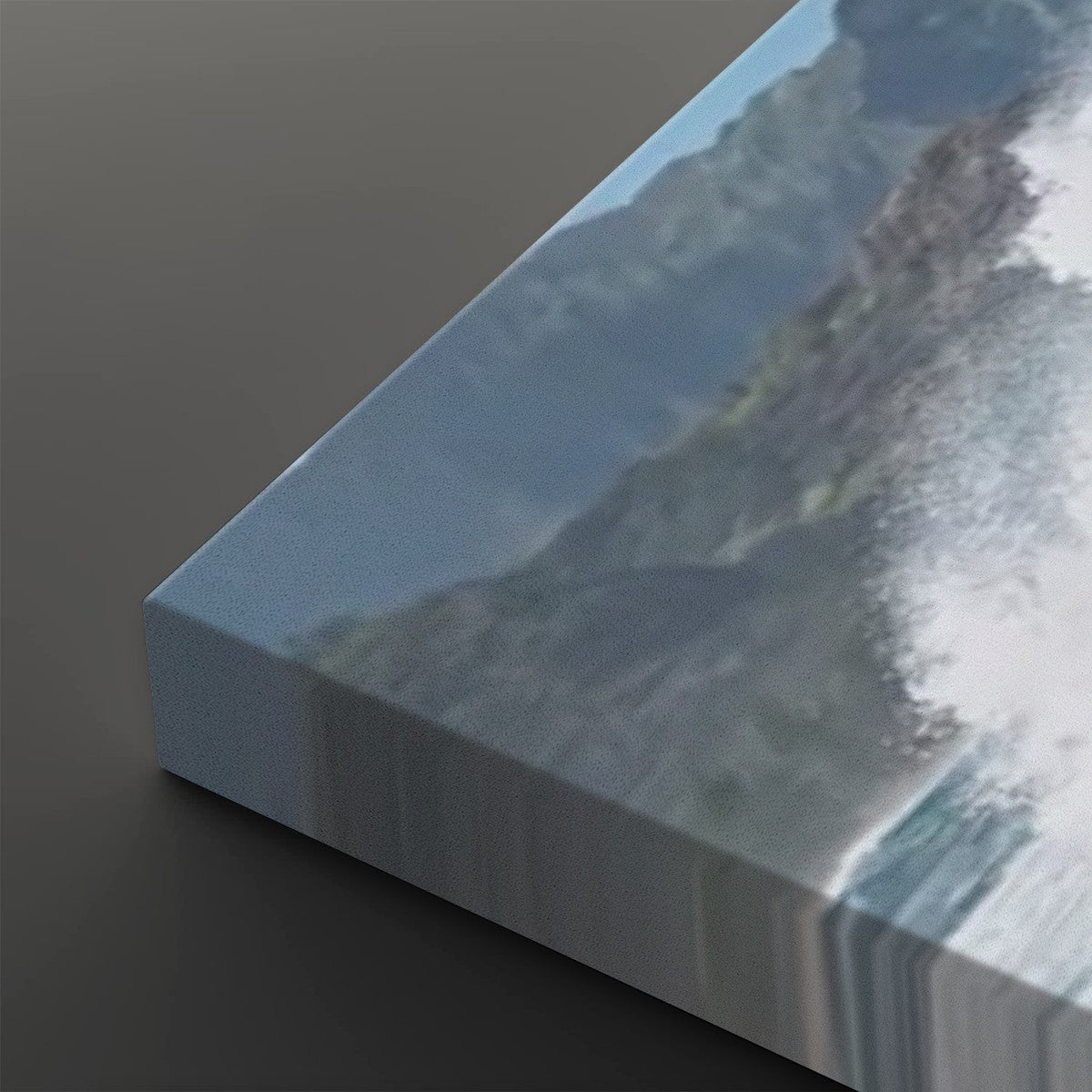 A close-up of a canvas wall art print showing an abstract coastal or mountain scene with blue-gray tones and white surf, photographed at an angle on a dark gray background.