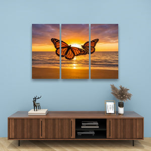 A three-panel canvas wall art set depicting two orange monarch butterflies over a beach sunset, displayed on a light blue wall above a wooden console table with decorative items.