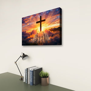 A rectangular wall art canvas showing a dark cross silhouette against a dramatic sunset sky with clouds and light rays, displayed on a light wall above a desk with books, a lamp, and a small plant.