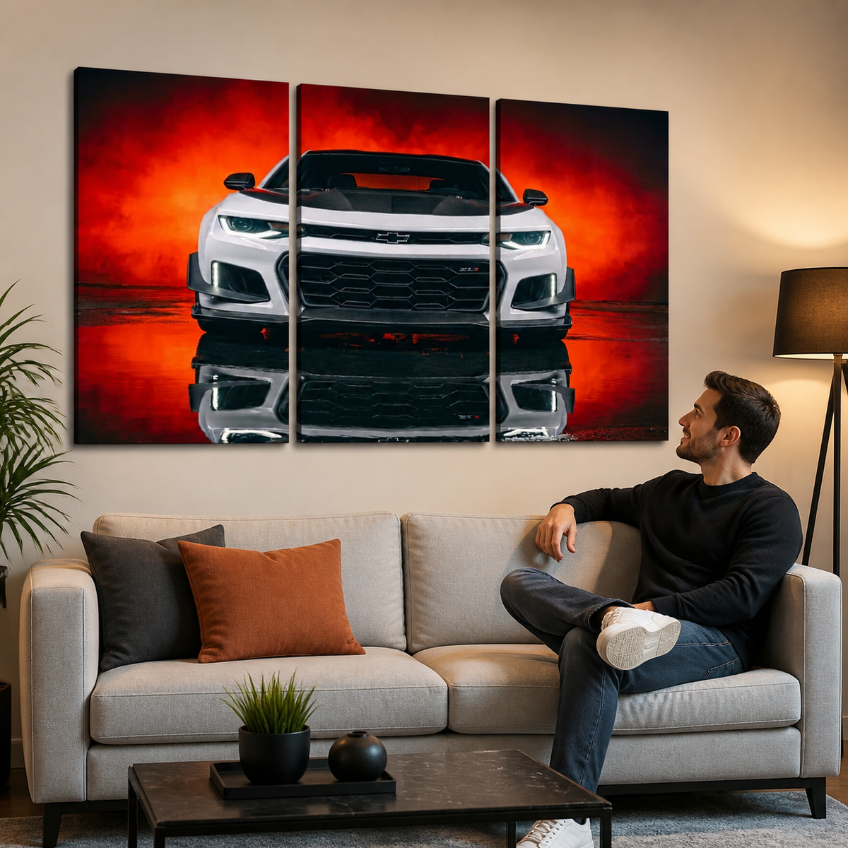 A man sits on a light beige sofa in a modern living room, looking at a three-panel canvas wall art print depicting a white Camaro  sports car against a dramatic red and orange smoky background with a reflection effect.