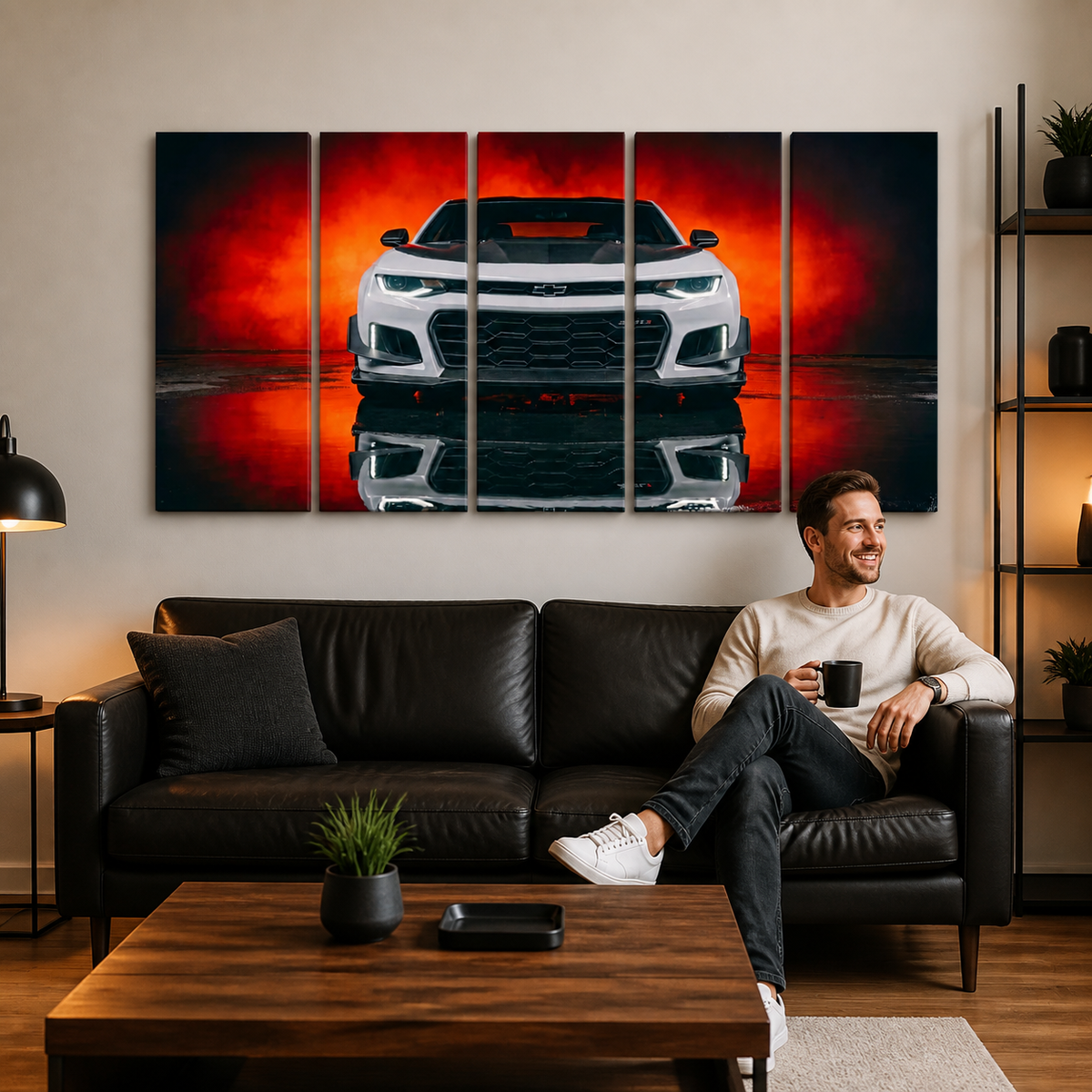A man sits on a black leather sofa in a modern living room, holding a black mug and smiling at a large five-panel canvas wall art print depicting a white Camaro sports car against a dramatic red and orange background.