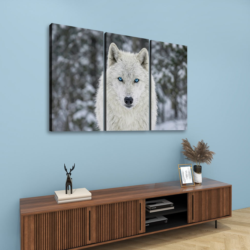 A three-panel canvas wall art set depicting a close-up portrait of a white wolf with blue eyes in a snowy outdoor setting, displayed on a light blue wall above a wooden console table with decorative items.