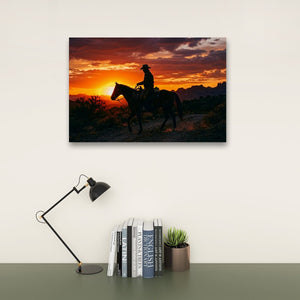 A rectangular wall art print showing a silhouetted cowboy riding a horse at sunset, displayed above a desk with a black lamp, books, and a small succulent on a light wall.