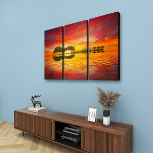 Three-panel canvas wall art depicting a guitar silhouette against a vivid red, orange, and yellow sunset, displayed above a wooden console table in a modern room setting.