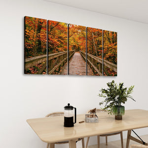 A five-panel canvas wall art set depicting a wooden boardwalk bridge over an autumn forest with vibrant orange and red foliage, displayed on a white wall above a dining table with a vase of greenery and a French press.