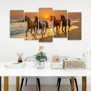 A 5-panel canvas wall art set depicting four horses walking on a beach at sunset, displayed on a white wall above a styled white dining table with books, a vase, and a basket.