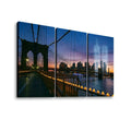 A three-panel canvas wall art print depicting the Brooklyn Bridge at dusk with the New York City skyline, including the Two Traqde Center Towers, over water. The left panel shows the bridge and walkway with warm lights, while the right panels show the city skyline under a purple and orange sunset sky.