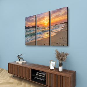 A three-panel canvas wall art set depicting a beach sunset with orange and pink sky, waves, and sand, displayed above a wooden console in a styled room with a blue wall.