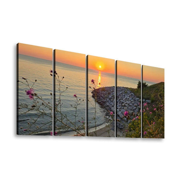 A five-panel split canvas wall art print depicting a coastal sunset over calm water, with a rocky shoreline, greenery, and pink wildflowers in the foreground, shown against a plain white background.