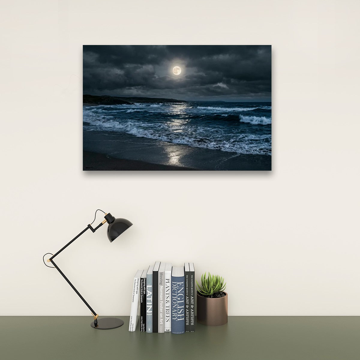 A rectangular wall art print showing a dark ocean shoreline under a cloudy night sky with a bright full moon, displayed above a desk with a black lamp, books, and a small potted succulent.
