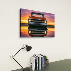 A rectangular wall art print showing a front-facing  1970 Chevrolet C10 truck reflected in water under a vivid sunset sky, displayed on a light wall above a desk with books, a small plant, and a black desk lamp.