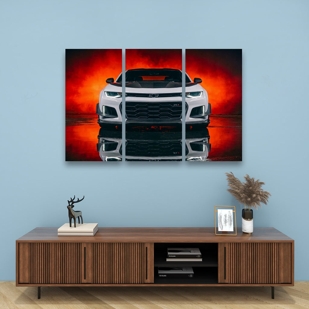 A three-panel canvas wall art print depicting a white Camaro sports car against a dramatic red and orange smoky background, displayed on a light blue wall above a wooden console table with decorative items.
