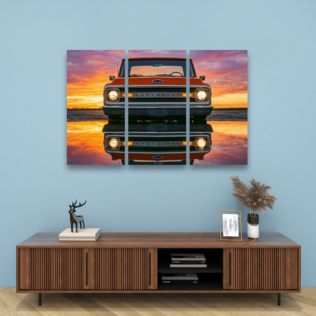 A three-panel canvas wall art print depicting a vintage orange pickup truck facing forward with its reflection on water under a pink, orange, and purple sunset sky, displayed on a light blue wall above a wooden console table.