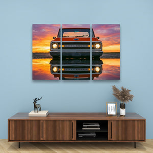 A three-panel canvas wall art print depicting a vintage orange pickup truck facing forward with its reflection on water under a pink, orange, and purple sunset sky, displayed on a light blue wall above a wooden console table.