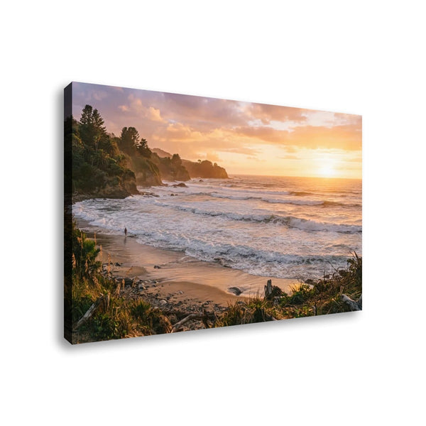 A rectangular canvas wall art print showing a coastal beach at sunset, photographed against a plain white background. The artwork depicts ocean waves, sandy shore, rocky cliffs, and trees under a glowing orange and pink sky.