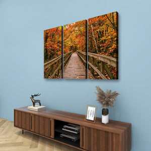 A three-panel canvas wall art set depicting a wooden boardwalk bridge over autumn-colored trees, displayed on a light blue wall above a wooden console table with decorative items.
