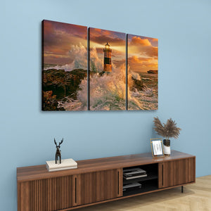 A three-panel canvas wall art set depicting a dramatic lighthouse scene with crashing waves and a golden sunset, displayed above a wooden console table against a light blue wall.