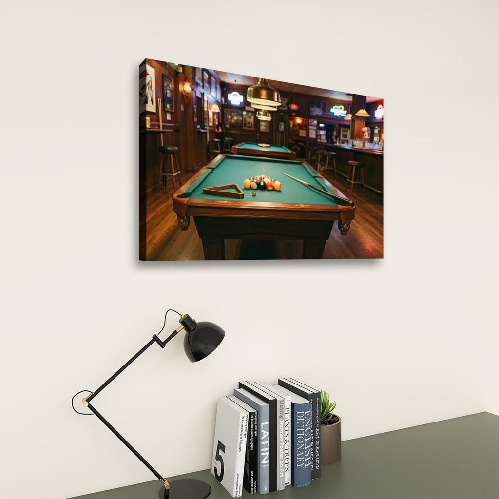 A rectangular wall art print showing a pool table in a dimly lit bar hangs on a light wall above a desk with a black lamp and books.