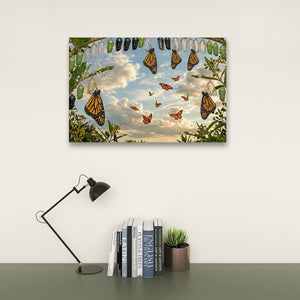 A framed wall art print depicting monarch butterflies flying through a blue sky with clouds, hanging leaves, and pupae, displayed above a desk with a black lamp, books, and a small potted succulent.