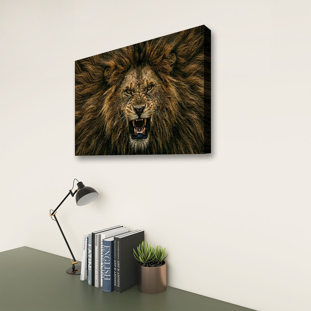 A rectangular wall art print showing a roaring lion's face, displayed above a desk with a lamp, books, and a small plant in a styled interior setting.