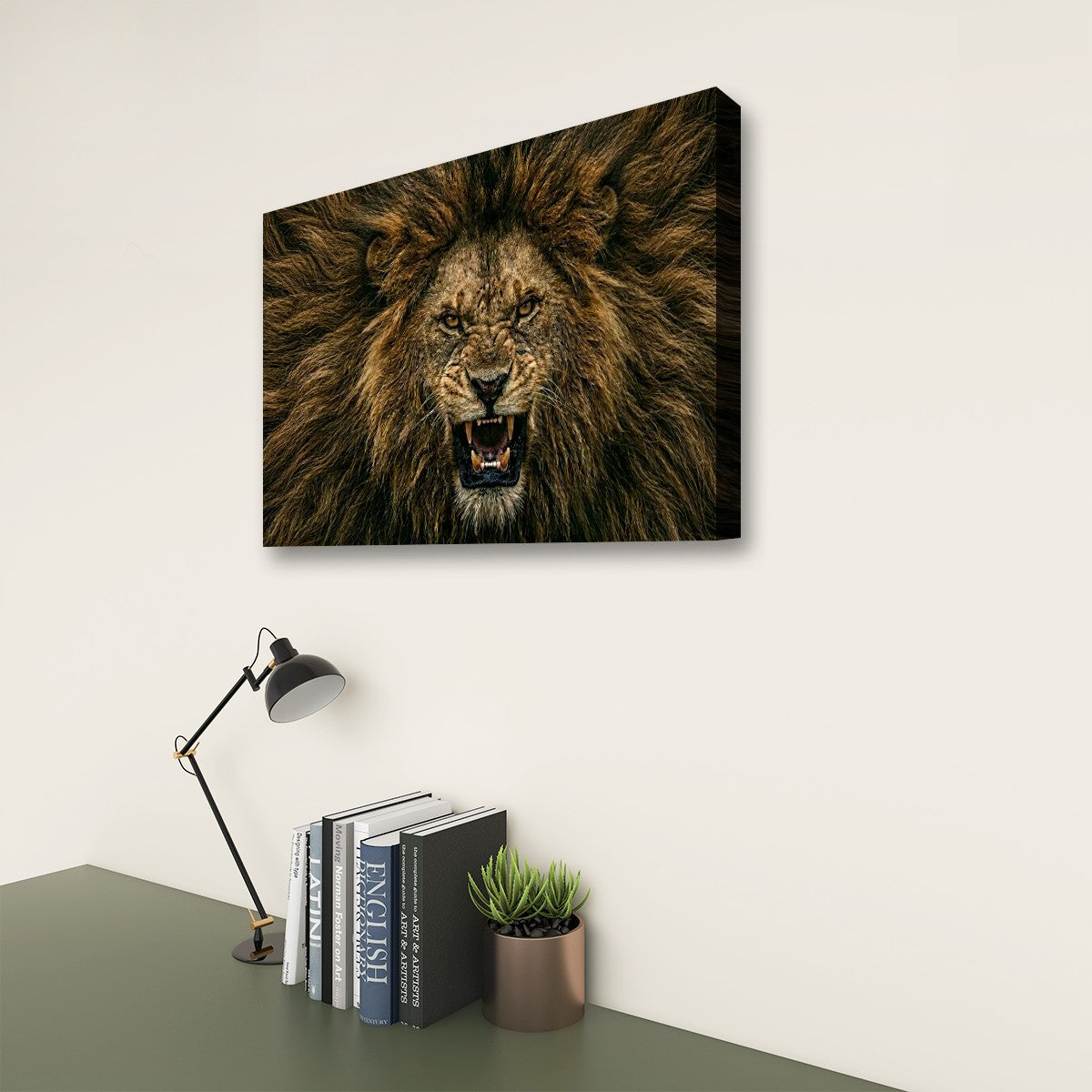 A rectangular wall art print showing a roaring lion's face, displayed above a desk with a lamp, books, and a small plant in a styled interior setting.