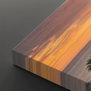 A close-up of a rectangular canvas or panel with a sunset-like abstract print, showing warm orange and yellow streaks over muted brown, gray, and purple tones, photographed on a dark gray background.