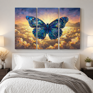 A three-panel canvas wall art set depicting a large blue butterfly over a glowing sunset sky and clouds, displayed above a neatly made bed in a modern bedroom.