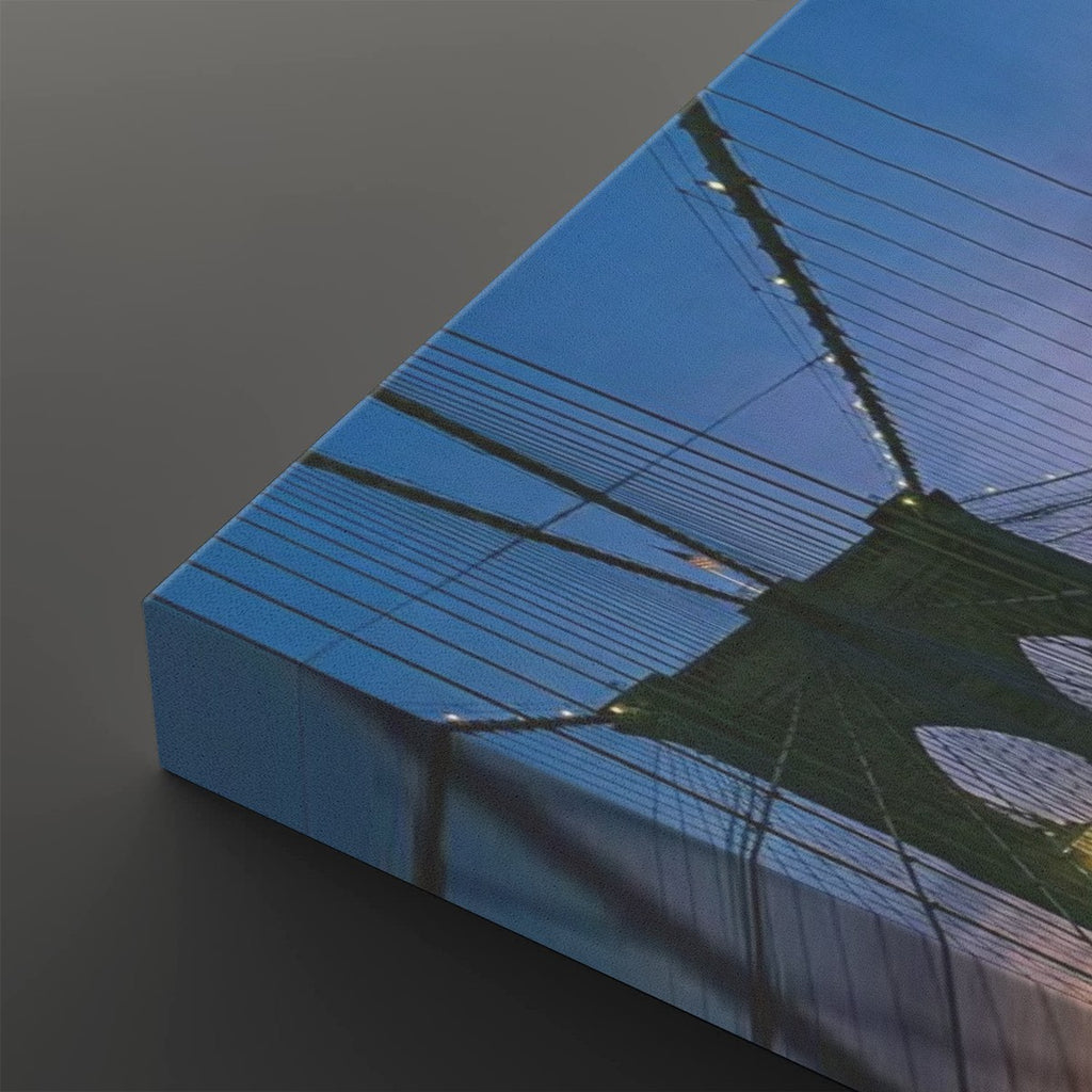 A close-up of a rectangular canvas print showing an upward view of the Brooklyn Bridge, with blue sky and bridge cables reflected on the gallery edge, set against a dark gray background.