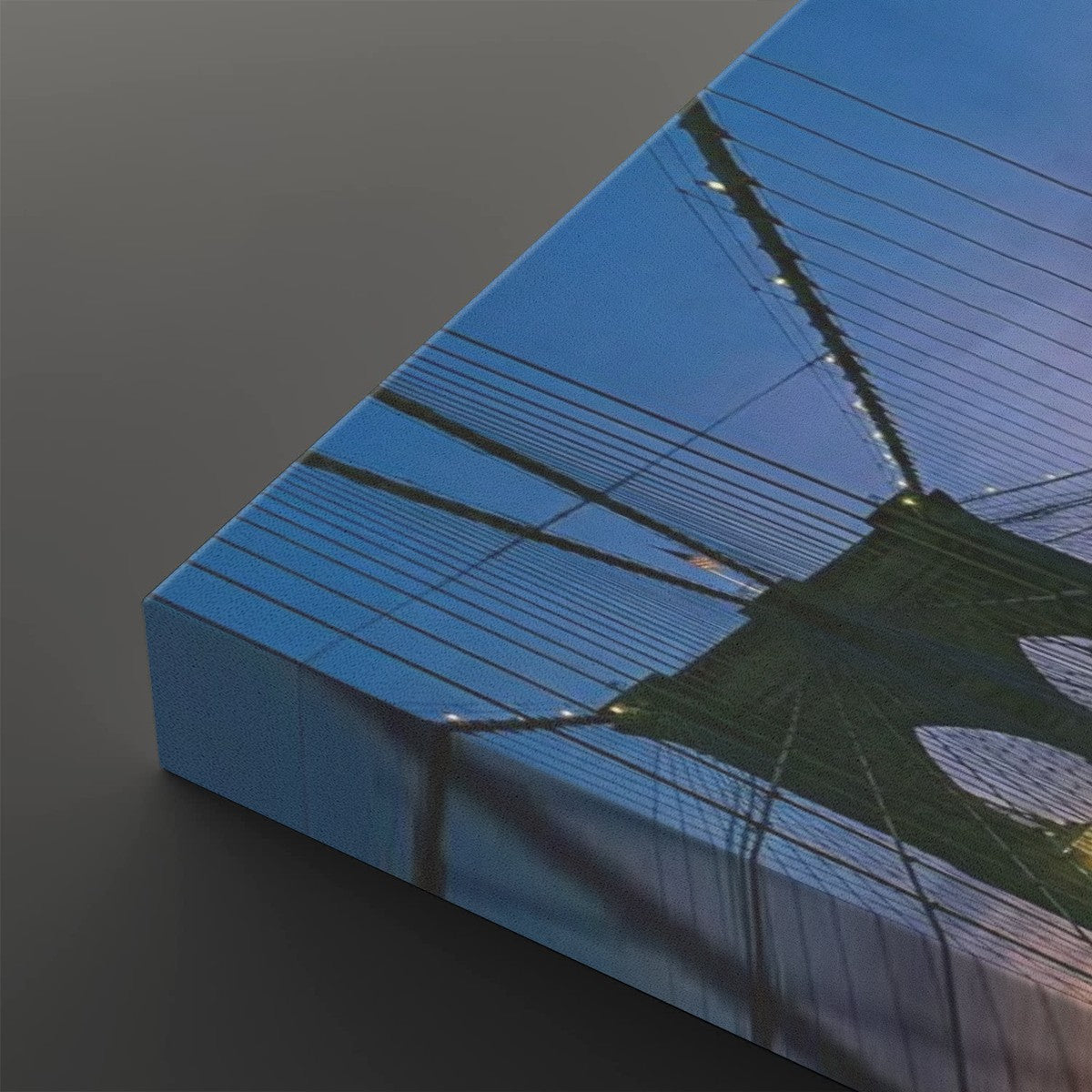 A close-up of a rectangular canvas print showing an upward view of the Brooklyn Bridge, with blue sky and bridge cables reflected on the gallery edge, set against a dark gray background.