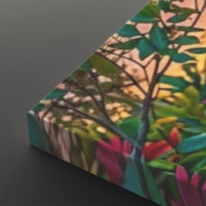 A close-up angled view of a rectangular canvas or print featuring a colorful blurred botanical scene with green leaves, pink and red flowers, and warm yellow-orange light, set against a dark gray background.