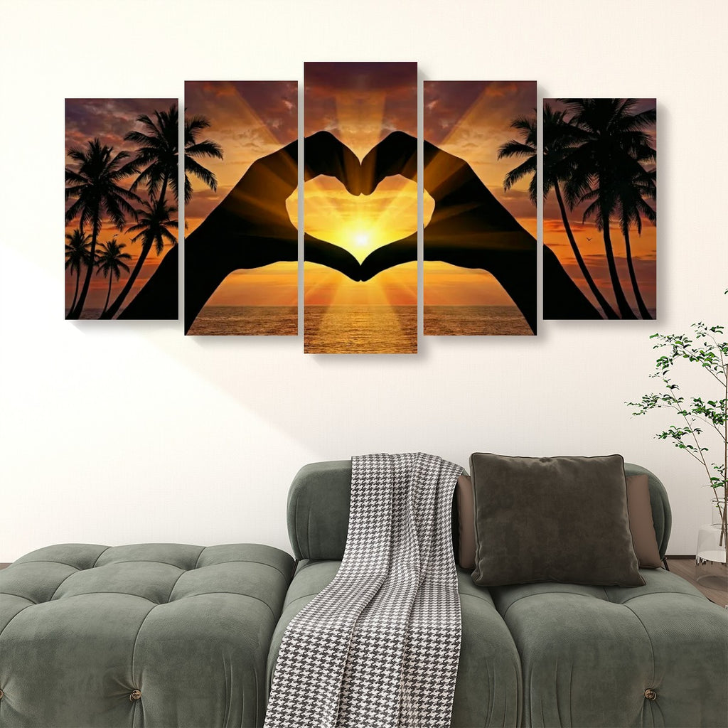 A five-panel canvas wall art set hanging above a green sofa in a living room. The artwork shows a sunset over the ocean with palm tree silhouettes and two large hand silhouettes forming a heart shape in the center.
