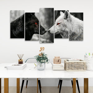 A 5-panel canvas wall art set depicting a black wolf and a white wolf facing each other, with glowing red eyes, displayed on a white wall above a modern white dining table with decorative items.