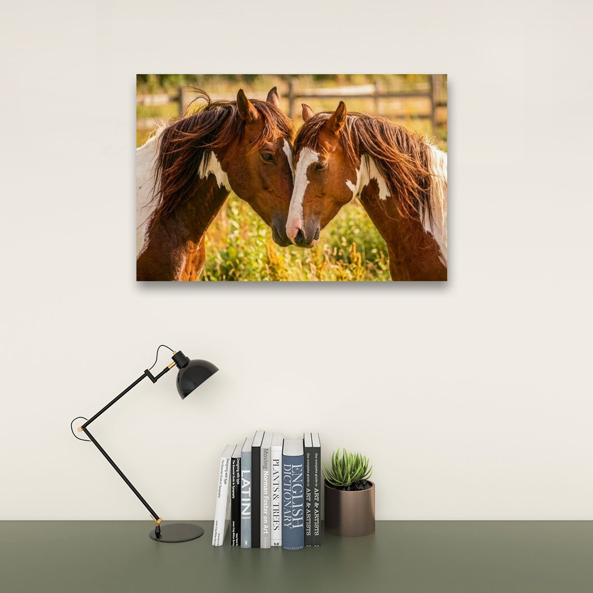 A rectangular wall art print showing two brown horses facing each other in a sunlit field, displayed above a desk with a black lamp, books, and a small succulent against an off-white wall.
