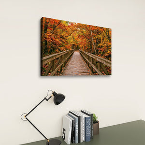 A rectangular wall art print showing a wooden boardwalk bridge over autumn-colored trees, displayed above a desk with a black lamp and books against a light wall.
