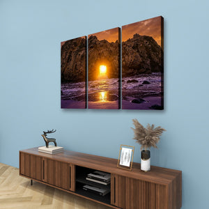 A three-panel canvas wall art set depicting a dramatic rocky coastline at sunset, displayed above a wooden console table against a light blue wall.