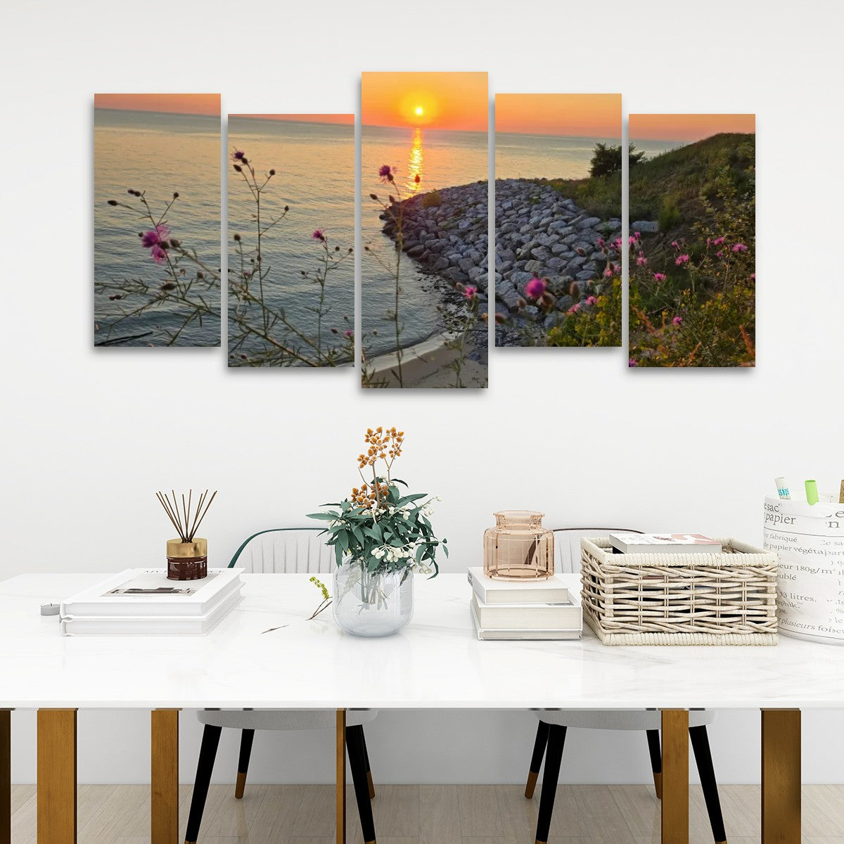 A five-panel canvas wall art set depicting a sunset over a rocky shoreline with wildflowers, displayed above a white dining table in a modern room setting.