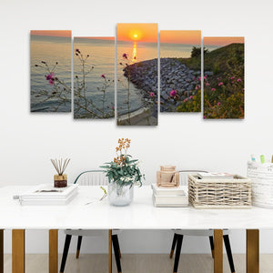 A five-panel canvas wall art set depicting a sunset over a rocky shoreline with wildflowers, displayed above a white dining table in a modern room setting.
