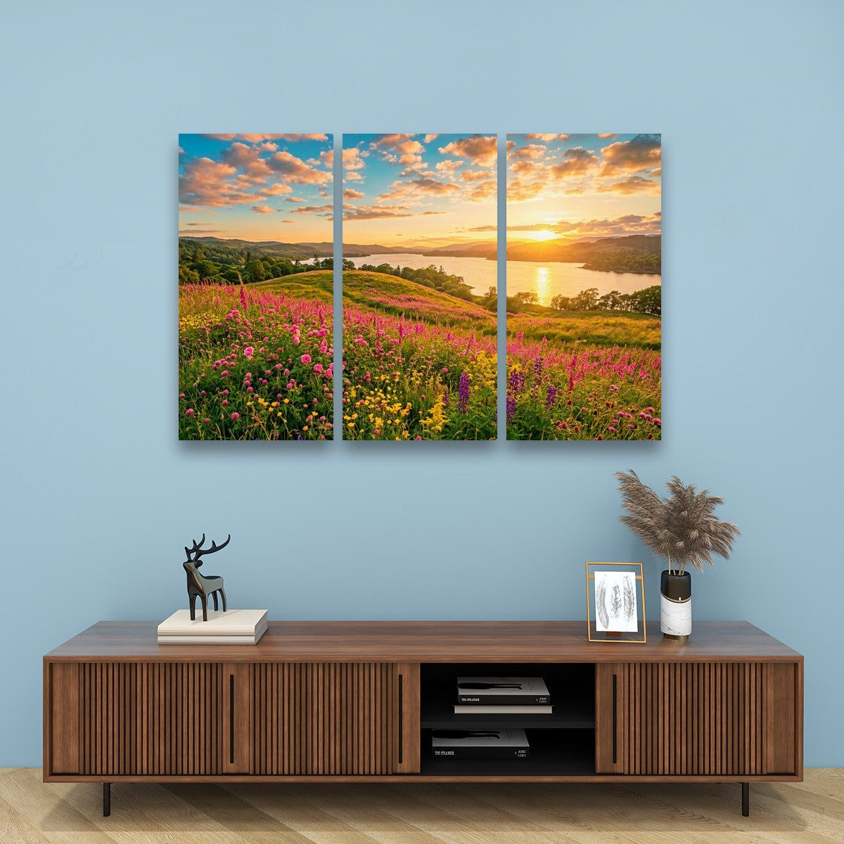 A three-panel canvas wall art set depicting a vibrant wildflower meadow at sunset by a lake, displayed on a light blue wall above a wooden console table with decorative items.