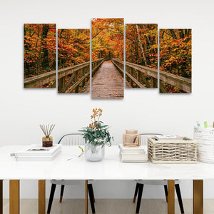 A five-panel canvas wall art set depicting a wooden boardwalk bridge over autumn foliage, displayed on a white wall above a styled dining table with books, a basket, and a vase.