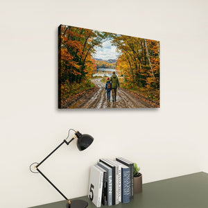 A rectangular wall art print showing two people walking on a dirt road surrounded by autumn trees, displayed above a desk with a black lamp and books against a light wall.