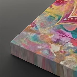 A close-up angled view of a rectangular canvas print showing a colorful abstract painted design with pink, purple, yellow, teal, and cream tones on a dark gray background.